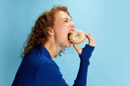Foto de Retrato de chica rizada joven en azul superior emocional comer donut contra fondo de estudio azul. Dulce diente. Concepto de emociones humanas, cultura juvenil, moda, estilo de vida, comida - Imagen libre de derechos