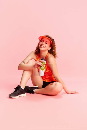 Foto de Retrato de una joven sonriente en ropa deportiva sentada en el suelo y bebiendo agua después de entrenar sobre el fondo rosa del estudio. Concepto de emociones humanas, cultura juvenil, moda, estilo de vida deportivo - Imagen libre de derechos