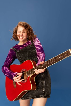 Téléchargez les photos : Portrait de jeune fille heureuse, souriante et émotionnelle jouant de la guitare, s'amusant sur fond de studio bleu. Concept d'émotions humaines, culture des jeunes, mode, style de vie créatif - en image libre de droit