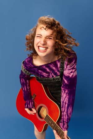 Photo for Portrait of happy, smiling, young, emotional girl playing guitar, having fun against blue studio background. Concept of human emotions, youth culture, fashion, creative lifestyle - Royalty Free Image