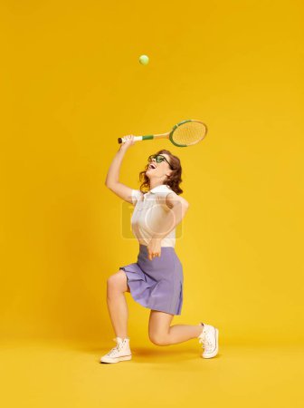 Téléchargez les photos : Portrait de jeune fille active et jolie en tenue de sport, jouant au tennis en mouvement frappant la balle avec raquette sur fond de studio jaune. Concept de sport, mode de vie active, émotions, mode, passe-temps - en image libre de droit