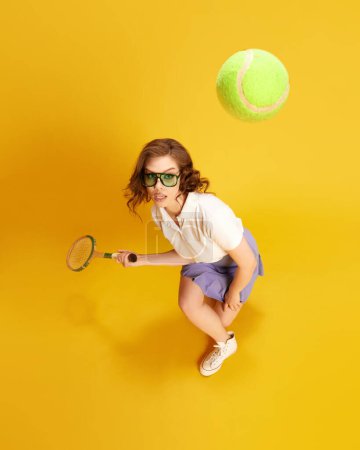 Foto de Retrato de vista superior de la joven chica concentrada, hermosa jugadora de tenis en movimiento, golpeando la pelota con raqueta contra el fondo amarillo del estudio. Concepto de deporte, estilo de vida activo, emociones, moda, hobby - Imagen libre de derechos