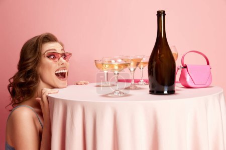 Foto de Retrato de una joven hermosa mujer asomándose a la mesa servida con champán sobre fondo de estudio rosa. Emoción y felicidad. Concepto de emociones, celebración, fiesta, moda - Imagen libre de derechos