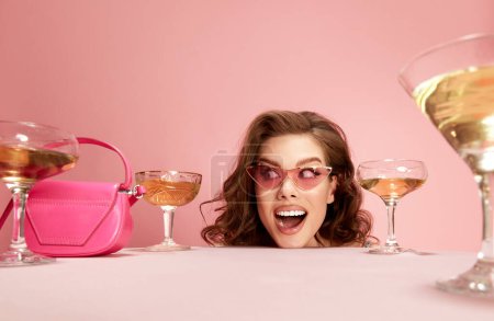 Foto de Retrato de una joven hermosa mujer asomándose a la mesa servida con champán sobre fondo de estudio rosa. Encantador, estado de ánimo positivo. Concepto de emociones, celebración, fiesta, moda - Imagen libre de derechos