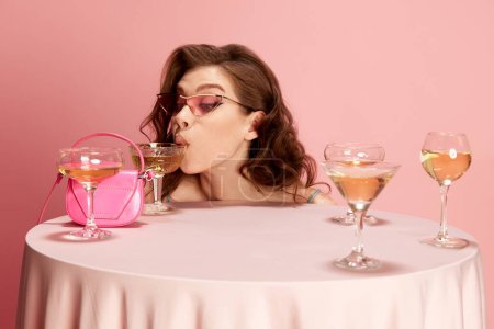 Foto de Retrato de una joven hermosa morena asomándose a la mesa servida con champán sobre fondo de estudio rosa. Bebiendo. Concepto de emociones, celebración, fiesta, moda - Imagen libre de derechos