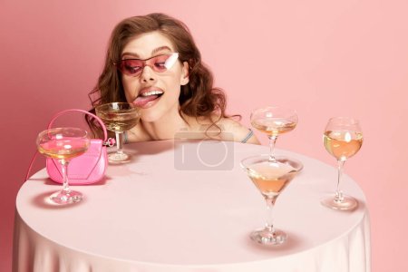 Foto de Retrato de una joven hermosa mujer asomándose a la mesa servida con champán sobre fondo de estudio rosa. Es un humor loco. Concepto de emociones, celebración, fiesta, moda, belleza - Imagen libre de derechos