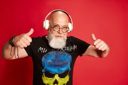 Photo for Portrait fp mature bearded, bald man with positive facial expression, listening to music in headphones against red studio background. Concept of human emotions, lifestyle, male fashion. Rock-n-roll - Royalty Free Image