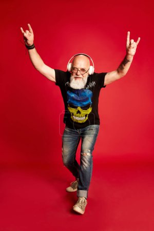 Photo for Rock-n-roll. Full-length portrait emotional, mature, bearded man with tattoos, listening to music in headphones against red studio background. Concept of human emotions, lifestyle, male fashion - Royalty Free Image
