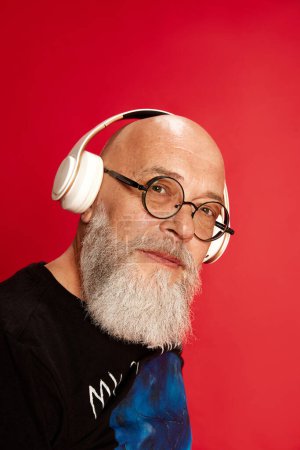 Photo for Close-up portrait of mature, bearded, bald man in glasses, listening to music in headphones and looking at camera against red studio background. Concept of human emotions, lifestyle, male fashion - Royalty Free Image