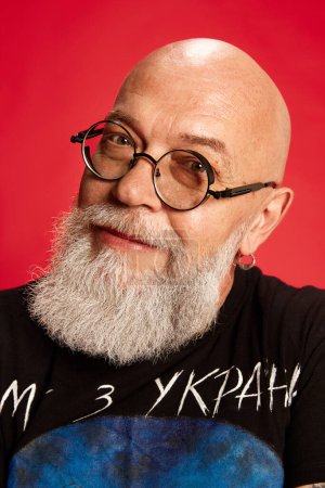 Photo for Portrait of bearded, mature, bald man in glasses and casual stylish clothes posing,m looking at camera and smiling against red studio background. Concept of human emotions, lifestyle, male fashion - Royalty Free Image