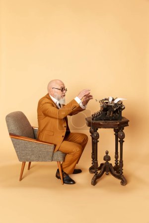Photo for Portrait of bearded, mature, bald man in elegant, classical suit sitting in front of typewriter and working against studio background. Concept of human emotions, lifestyle, business, art, fashion - Royalty Free Image