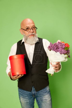 Téléchargez les photos : Portrait d'homme mûr, barbu et chauve en vêtements et lunettes élégants, debout avec des fleurs sur fond de studio vert. Concept d'émotions humaines, style de vie, mode masculine, célébration, salutations - en image libre de droit