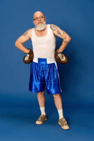 Téléchargez les photos : Portrait d'homme chauve, barbu et mature en tenue de sport et lunettes, avec corps tatoué en gants de boxe vintage sur fond de studio bleu. Concept d'émotions, sport, mode de vie active, mode sportive - en image libre de droit