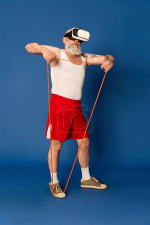 Téléchargez les photos : Portrait d'un homme mûr et barbu en tenue de sport et lunettes vr avec des extenseurs de fitness sur fond de studio bleu. Formation en ligne. Concept de sport, mode de vie actif, santé - en image libre de droit