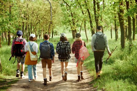 Foto de Jóvenes activos y deportivos, amigos haciendo senderismo con mochilas en el bosque en un cálido día de primavera, caminando por el camino, disfrutando de paisajes. Concepto de estilo de vida activo, naturaleza, deporte y hobby, amistad - Imagen libre de derechos