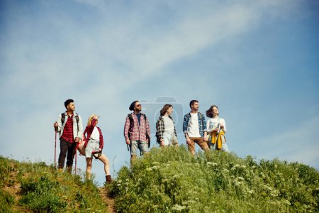 Foto de Grupo de amigos caminando en el prado en un día cálido, soleado y de verano. Jóvenes de pie en la colina y disfrutando de la naturaleza, el paisaje. Concepto de estilo de vida activo, naturaleza, deporte y hobby, amistad - Imagen libre de derechos