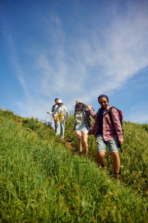 Foto de Grupo de amigos, jóvenes caminando en el prado, bajando la colina en un día cálido y soleado. Hombres ayudando a las mujeres. Concepto de estilo de vida activo, naturaleza, deporte y hobby, amistad - Imagen libre de derechos