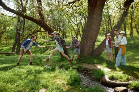 Foto de Jóvenes, hombres y mujeres, amigos haciendo senderismo, caminando por el bosque, saltando sobre el río en un día cálido. Divirtiéndose al aire libre. Concepto de estilo de vida activo, naturaleza, deporte y hobby, amistad - Imagen libre de derechos