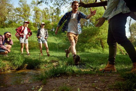 Foto de Grupo de amigos, jóvenes activos haciendo senderismo, caminando por el bosque, saltando sobre el río y divirtiéndose al aire libre en el cálido día de primavera. Concepto de estilo de vida activo, naturaleza, deporte y hobby, amistad - Imagen libre de derechos