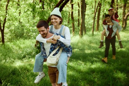 Foto de Dos parejas jóvenes, hombres y mujeres caminando por el bosque, haciendo senderismo en un cálido día soleado. Chicas sentadas sobre espaldas de chicos. Concepto de estilo de vida activo, naturaleza, deporte y hobby, amistad, ocio y diversión - Imagen libre de derechos