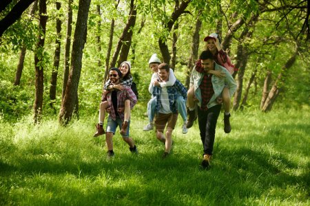 Foto de Grupo de jóvenes, amigos caminando en el bosque, haciendo senderismo en un cálido día soleado. Chicas sentadas sobre espaldas de chicos. Concepto de estilo de vida activo, naturaleza, deporte y hobby, amistad, ocio y diversión - Imagen libre de derechos