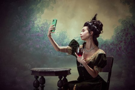 Téléchargez les photos : Portrait de jeune belle fille, princesse en robe élégante prenant selfie avec téléphone portable et boire du vin rouge sur fond vintage sombre. Concept d'histoire, art de la renaissance, comparaison des époques - en image libre de droit