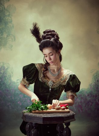 Téléchargez les photos : Portrait de jeune fille tendre, princesse élégante dans la cuisine élégante robe médiévale, couper les légumes sur fond sombre vintage. Concept d'histoire, art de la renaissance, comparaison des époques - en image libre de droit