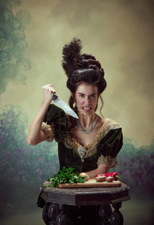 Téléchargez les photos : Portrait de jeune fille émotionnelle en colère, princesse médiévale tenant un couteau sur des légumes et montrant une irritation sur fond sombre vintage. Histoire, art de la renaissance, comparaison des époques concept - en image libre de droit