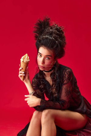 Téléchargez les photos : Portrait de jeune belle fille en élégante robe noire assise, mangeant de la crème glacée sur fond rouge. Concept d'histoire, art de la renaissance, comparaison des époques, nourriture, beauté, mode - en image libre de droit