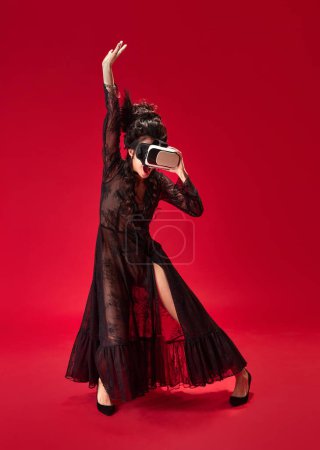 Foto de Retrato de larga duración de una joven en un elegante vestido negro con gafas VR y bailando sobre fondo rojo. Concepto de historia, arte renacentista, comparación de épocas, tecnologías modernas - Imagen libre de derechos