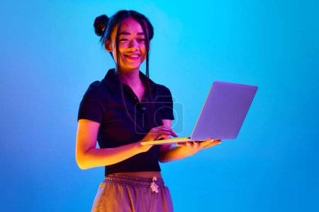 Téléchargez les photos : Portrait de jeune fille coréenne, étudiant debout avec ordinateur portable sur fond de studio bleu sous la lumière du néon. Concept d'émotions, expression faciale, jeunesse, mode de vie, éducation, entreprise, publicité - en image libre de droit