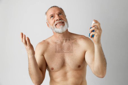 Photo for Portrait of handsome, smiling, mature man with gray hair and beard posing shirtless, applying with face toner against grey background. Concept of male beauty, face and skin care, daily procedures, age - Royalty Free Image