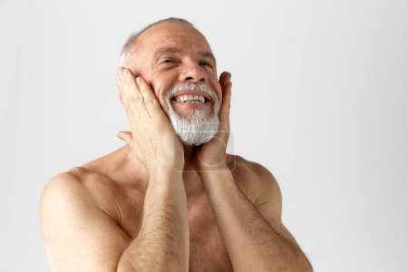 Photo for Portrait of handsome, mature, smiling man with gray-haired beard, applying face lotion against grey background. Concept of male beauty, face and skin care, daily procedures, age, cosmetology - Royalty Free Image
