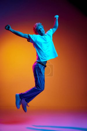 Téléchargez les photos : Portrait complet d'une mature en vêtements décontractés sautant sur fond de studio orange en néon bleu. Bonheur et succès. Concept d'émotions humaines, mode de vie, jeunesse, expression faciale - en image libre de droit
