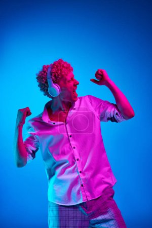 Photo for Portrait of emotive mature man in white shirt listening to music in headphones against blue studio background in pink neon light. Concept of human emotions, lifestyle, youth, facial expression - Royalty Free Image