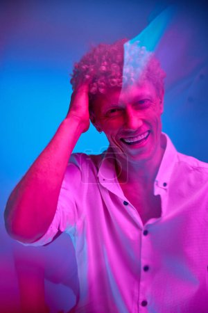 Foto de Retrato de hombre con el pelo rizado en la camisa blanca posando, sonriendo alegremente sobre fondo de estudio azul en luz de neón rosa. Concepto de emociones humanas, estilo de vida, juventud, expresión facial - Imagen libre de derechos