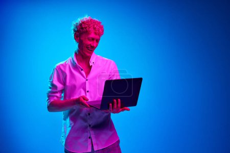 Foto de Retrato de hombre rizado en camisa blanca trabajando con portátil contra fondo de estudio azul en luz de neón rosa. Sonriendo, riendo. Concepto de emociones humanas, estilo de vida, negocios, expresión facial - Imagen libre de derechos