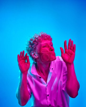 Photo for Portrait of man with curly hair, in white shirt, leaning on transparent glass against blue studio background in pink neon light. Funny face. Concept of human emotions, lifestyle, facial expression - Royalty Free Image
