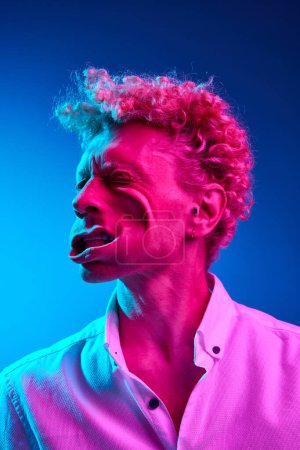 Photo for Funny image of man with curly hair posing with blowing wind on face against blue studio background in pink neon light. Concept of human emotions, lifestyle, youth, facial expression - Royalty Free Image