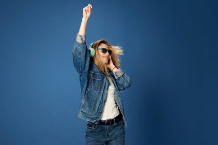 Photo for Portrait of young girl in jeans outfit listening to music in headphones, smiling, dancing against blue studio background. Concept of youth, human emotions, facial expression, fun, leisure - Royalty Free Image