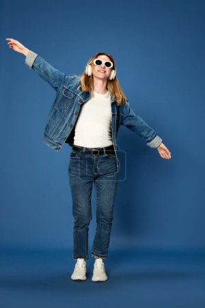 Foto de Retrato de larga duración de una joven vestida con jeans escuchando música en auriculares, sonriendo sobre fondo azul del estudio. ¡Joy! Concepto de juventud, emociones humanas, expresión facial - Imagen libre de derechos