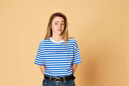 Photo for Portrait of young girl in striped shirt standing with doubtful face against yellow studio background. Misunderstanding. Concept of youth, human emotions, facial expression - Royalty Free Image