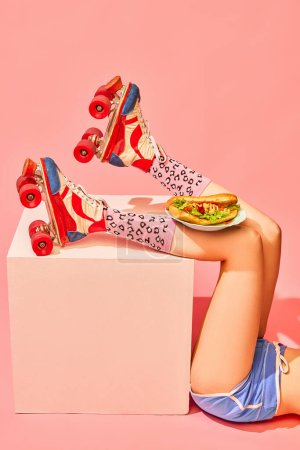 Foto de Patas femeninas delgadas en rodillos vintage con delicioso hot-dog sobre fondo rosa. Servicio de comida y entrega. Concepto de fotografía pop art, visión creativa, imaginación. Arte mínimo - Imagen libre de derechos