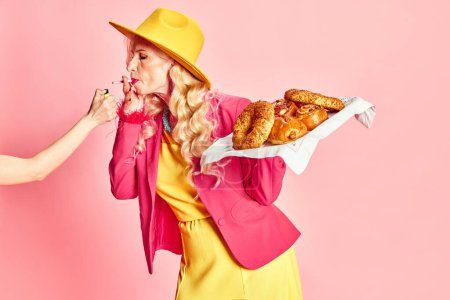 Photo for Portrait of attractive, stylish, senior woman in colorful clothes smoking cigarette and holding fresh sweet buns against pink studio background. Concept of beauty, fashion, human emotions, lifestyle - Royalty Free Image