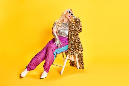 Photo for Portrait of stylish, beautiful senior woman in sportswear, heeled boots and animal print coat sitting against yellow studio background. Concept of beauty, fashion, human emotions, lifestyle. Ad - Royalty Free Image