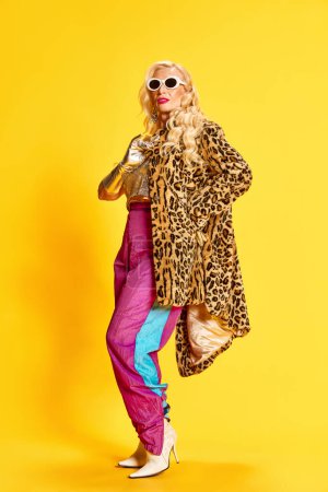Photo for Full-length portrait of stylish, beautiful senior woman in sports pants, heeled boots and animal print coat sitting against yellow studio background. Beauty, fashion, emotions, lifestyle concept. Ad - Royalty Free Image