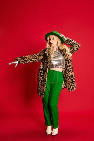 Photo for Portrait of beautiful senior lady, woman in stylish clothes, coat and green hat posing against red studio background. Birthday celebration. Concept of beauty, fashion, human emotions, lifestyle. Ad - Royalty Free Image
