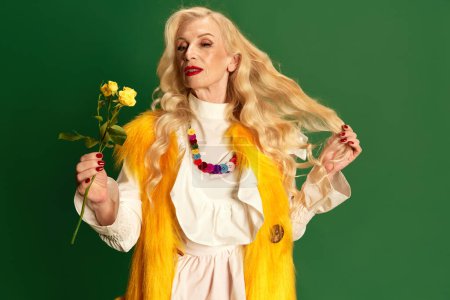 Téléchargez les photos : Portrait de belle dame avec de longs cheveux blonds et maquillage, femme âgée dans des vêtements élégants tenant des roses jaunes sur fond de studio vert. Concept de beauté, mode, émotions humaines, mode de vie - en image libre de droit