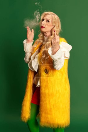 Photo for Portrait of cheeky senior woman smoking and showing rude gesture fish finger against green studio background. Expressive mood. Concept of beauty, fashion, human emotions, lifestyle. Ad - Royalty Free Image
