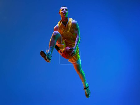 Téléchargez les photos : Jeune homme musclé, joueur de basket professionnel en mouvement, s'entraînant, sautant avec une balle contre fond de studio bleu sous la lumière du néon. Concept de sport professionnel, passe-temps, mode de vie sain, action - en image libre de droit